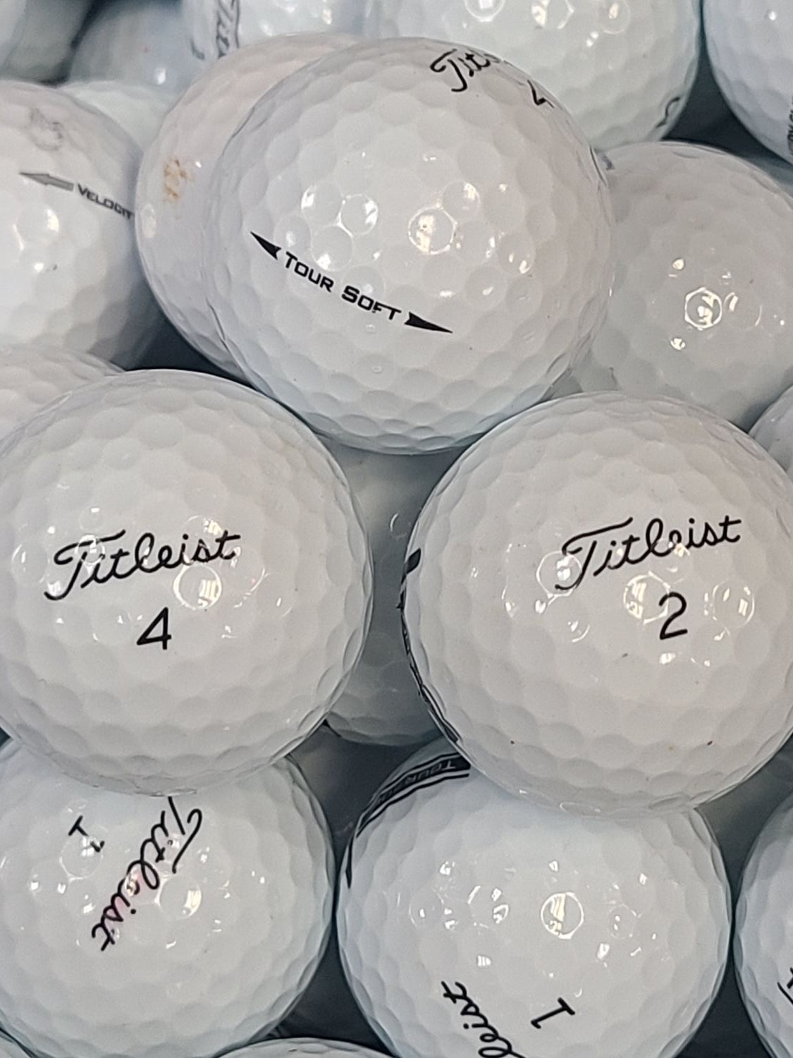 A close-up pile of white Titleist golf balls, some with black handwritten numbers and others showing the Titleist logo and TOUR SOFT text.